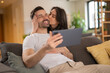 © Dexon Dee - Happy young couple sitting on a couch in a cozy living room. The woman hugs and kisses the man on the cheek while he holds a tablet. They both look joyful and connected, enjoying their time together.