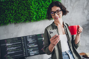 Brunette Female Programmer Working Remotely with a Smartphone and Coffee in a Modern Workspace with Computer Monitors