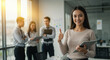 © schiers_images - A woman stands in an office, holding a tablet and giving a thumbs-up, while three people stand in the background, engaged in a discussion.