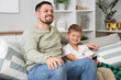© Pixel-Shot - Happy little son and his father watching TV on sofa at home