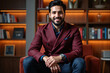 © Alex & M Studio - Confident South Asian businessman in burgundy double-breasted blazer sitting relaxed in modern office with illuminated bookshelf backdrop, showcasing professional style