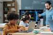 © pressmaster - Diverse group of children engaging in a classroom session with laptops while teacher explaining computer code in background. Focused young learners sitting attentively at desks