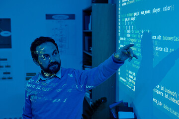 Man pointing at projected digital screen with coding and software development visuals. Office space includes various desk items and shelves in background