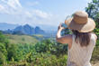 © Vladimir Razgulyaev - A woman in a Straw hat takes pictures of nature on her phone, rear view, from a viewing point. A popular landscape destination in Thailand is Khao Sok Park, mountains and rainforest panorama.