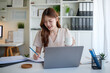 © wattana - Smiling young woman working on laptop at home office, taking notes in a notebook.