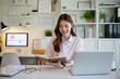 © wattana - Smiling young woman working on laptop at home office, taking notes in a notebook.