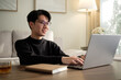 © wattana - Young man in glasses typing on a laptop while seated on a sofa in a cozy living room with a tea cup on the table.