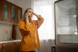 © Grustock - Frustrated woman in orange shirt talking on mobile phone, gesturing towards empty refrigerator in bright kitchen, reporting malfunction to customer service
