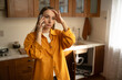 © Grustock - A woman in a yellow shirt stands in her kitchen while discussing urgent plumbing issues with a repair service regarding a leaky sink that needs immediate attention.