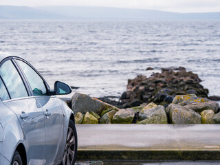 Wall Mural - A car is parked on the side of a road near the ocean. The car is silver. The ocean is calm and the sky is cloudy. Mountains in the background. Travel to nature concept.