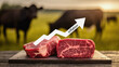 © jocaja - Dos filetes de carne vacuna cruda sobre tabla de madera, con gráfico de inflación al alza y vacas pastando al fondo.
