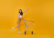 © NaMong Productions - Full length happy excited Asian teen woman pushing empty shopping cart or shopping trolley pointing finger promotion supermarket isolated on yellow background.
