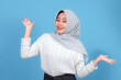 © Saddha Visual - Smiling young woman in hijab making a playful hand gesture with both hands raised, wearing a white knitted sweater against a blue background, expressing cheerful energy.