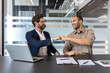 © Liubomir - Two men, likely colleagues, bump fists in a modern office environment. A laptop and documents are on the table, indicating a business meeting or discussion.