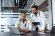 © Liubomir - Two doctors in white coats are discussing something on a laptop, a clipboard, and a tablet in a modern office setting.