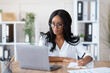© Prostock-studio - Happy black young business lady working on laptop at office. Focused African American woman taking notes during online work meeting or webinar, writing down important information