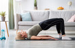© Prostock-studio - Stay at home fitness. Strong mature woman doing half bridge yoga pose, strengthening her abs muscles indoors, copy space. Fit senior woman working out in living room during covid isolation
