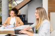 © Moon Safari - Focused businesswoman working online over laptop and female colleague reviewing reports at desk in office