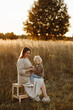 © Kaplitskaya Love - Front view of a young caucasian mother in a light dress holding her smiling toddler daughter on her lap in a wheat field at sunset. Concept parenting, love,autumn, family bonding, and rural lifestyle.