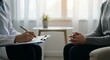 © Grosir - Therapist Writes Notes During Session with Patient Clasped Hands Illuminated by Soft Diffused Light in Modern Office