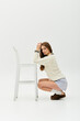 © LIGHTFIELD STUDIOS - Young beautiful woman kneeling beside a white stool in a bright, minimalist studio space