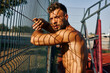 © djile - Muscular athlete resting against a metal fence on a sports field enjoying the sun and fresh air after intense workout