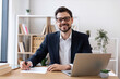 © sofiko14 - Smiling young Caucasian adult male in formal suit working at desk with laptop and papers. Professional, productive, and focused business setting showcasing modern office atmosphere.