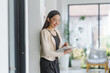 © amnaj - Young Asian businesswoman smiling and writing notes on a clipboard in a modern office, enjoying her productive workday