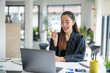 © amnaj - Young Asian businesswoman pointing at laptop while engaged in a video call, showcasing a modern office environment filled with professionalism