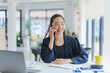 © amnaj - Young manager engaging in a phone conversation while working at her desk, surrounded by a laptop, documents, and a bright workspace