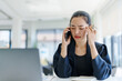 © amnaj - Asian manager feeling tired and frustrated after phone call with client, touching temples to relieve headache, sitting at desk with laptop and documents