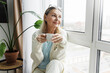 © shurkin_son - Retired woman of 65 enjoying coffee in morning admiring beauty of waking city through window of her living-room of multi-storey apartment block, holding white mug in hands sitting on windowsill