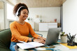 © Prostock-studio - People And Technology. Portrait of young smiling black woman in wireless headset using pc sitting at table on chair in living room, typing on keyboard. Cheerful lady browsing internet, surfing web