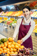 © JackF - Vegetable shop assistant working in greengrocery. Woman seller in sales area with tangerine in hands. Employee demonstrates supply of fruit. Local and imported farm vegetables and fruits
