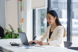 © amnaj - Young Asian businesswoman smiling and working using laptop computer and taking notes in modern office