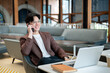 © bongkarn - Brown suit asian man with glasses is smiling while talking on phone, sitting with laptop in cafe.