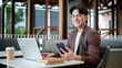 © bongkarn - Brown suit asian man with glasses is smiling while holding his phone sitting with laptop in the cafe