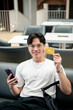 © bongkarn - Asian man wearing glasses in white tee holding phone and showing his credit card while sit on sofa.