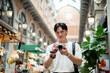 © bongkarn - Asian man wearing glasses looking ahead while holding camera in hallway of shopping mall or market