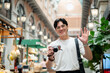 © bongkarn - Asian man wearing glasses waving a hand while holding a camera in hallway of shopping mall or market