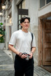 © bongkarn - Asian man wearing glasses in white tee looking ahead with camera in hand at shopping mall or market.