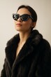 © FantasyLand86 - A stylish woman wearing sleek black cat-eye sunglasses and a luxurious dark faux fur jacket, standing confidently with her head tilted slightly, looking upwards.