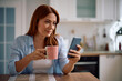 © Drazen - Happy woman texting on mobile phone while having cup of tea at home.