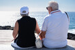 © luciano - Rear view of caucasian senior pensioner couple sitting face the sea enjoying freedom and vacation, two elderly people in retirement stay together in good harmony. Horizon over water