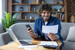 © Liubomir - A happy man examines a bill while using his smartphone and laptop, working on his finances from home.
