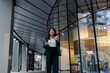 © Westend61 - Businesswoman walking outside a modern office building holding a laptop and coffee