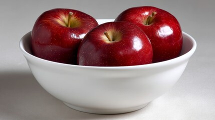 Aufkleber - Three vibrant red apples are closely packed in a smooth white bowl, illuminated by soft lighting that enhances their glossy texture and creates gentle reflections