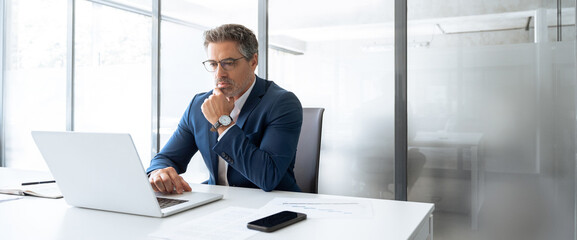Focused latin senior in suit financial expert analyst thinking on sale solution. Professional indian manager investor using pc laptop computer sitting at desk workplace in office. Banner, copy space