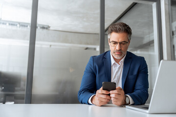 Busy mature ceo businessman in suit using phone for business work in office. Worried senior finance professional man working on cellphone telephone, pc laptop computer looking at screen. Copy space