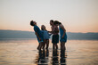 © qunica.com - A group of friends joyfully laughing while standing in the water under a colorful sunset, showcasing togetherness and happiness in picturesque surroundings.